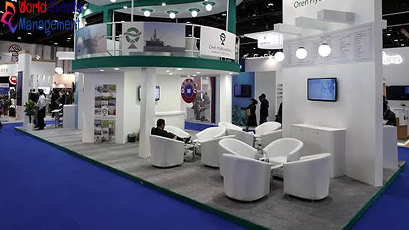 White and blue exhibition booth with meeting tables, digital screens, and overhead branding